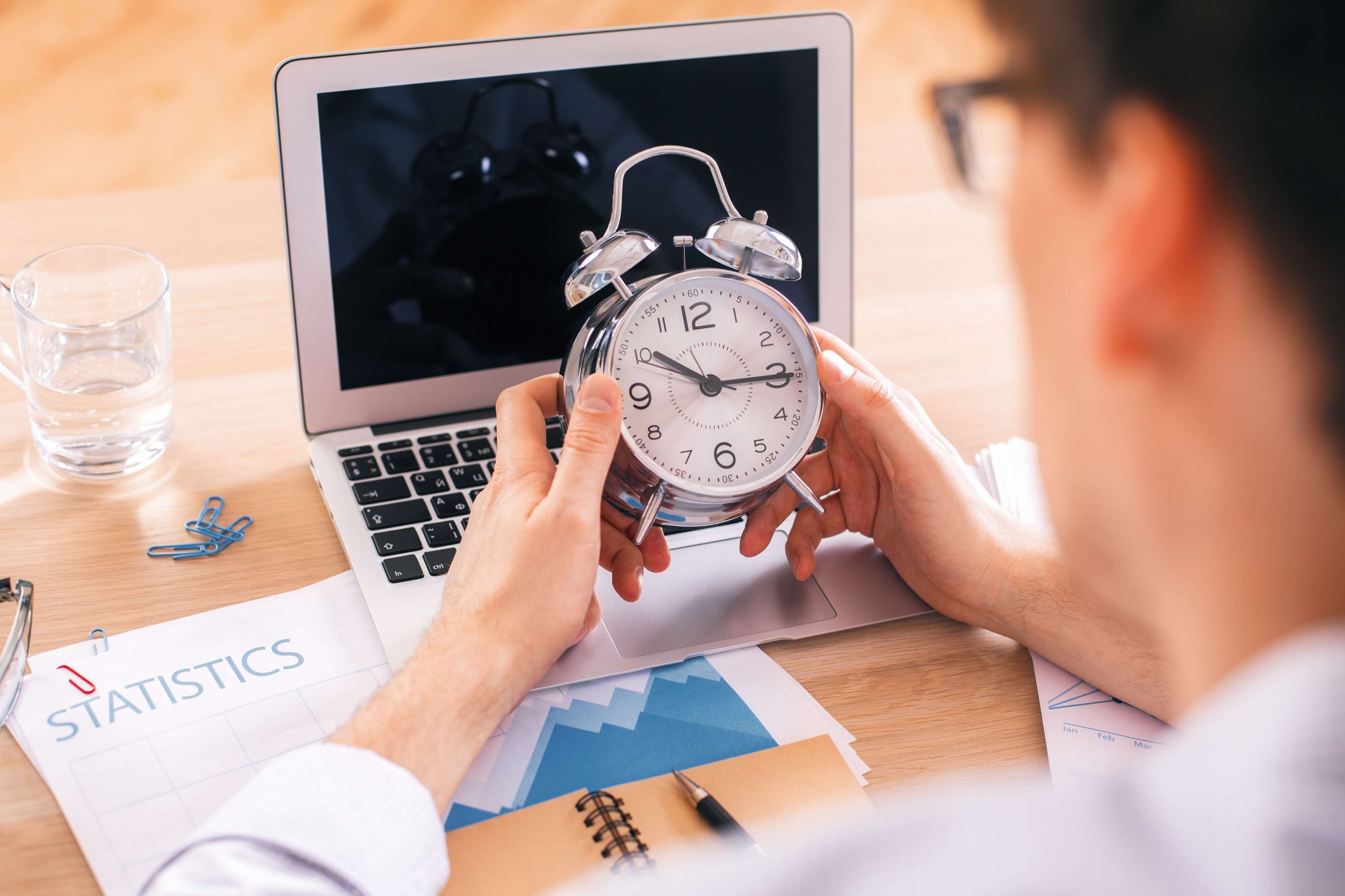 Male holding alarm clock over desktop with laptop, water glass and other items