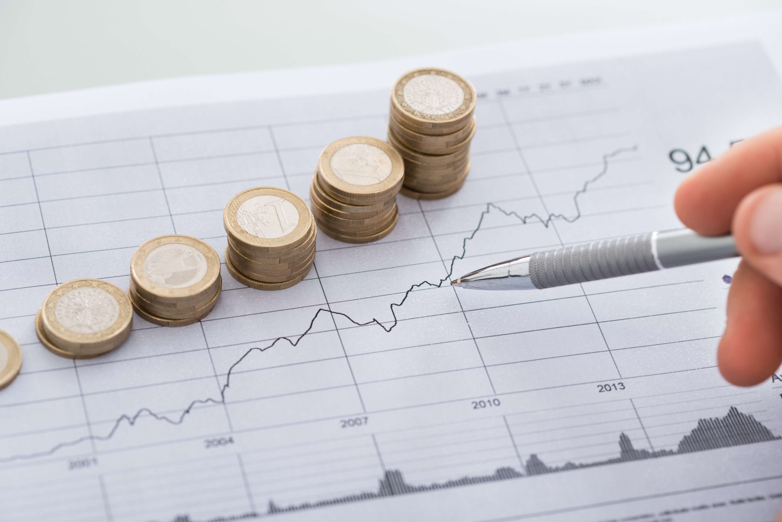 Hand With Pen Analyzing Line And Coin Graphs On Desk