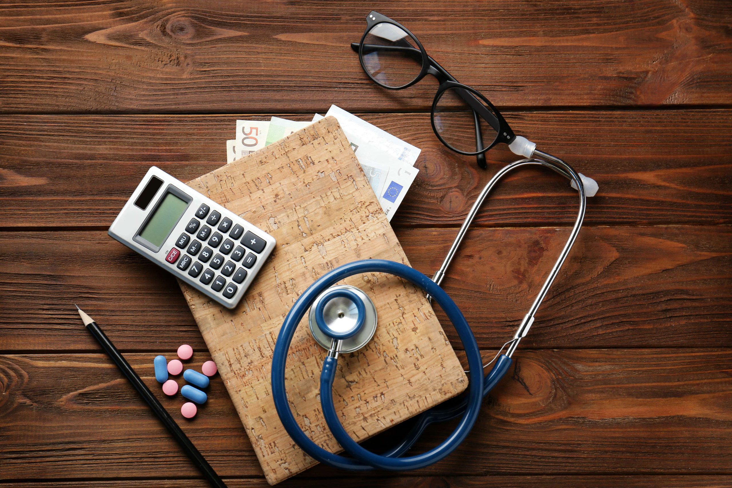 Medical concept. Notebook with money, calculator, stethoscope and pills on wooden table, top view