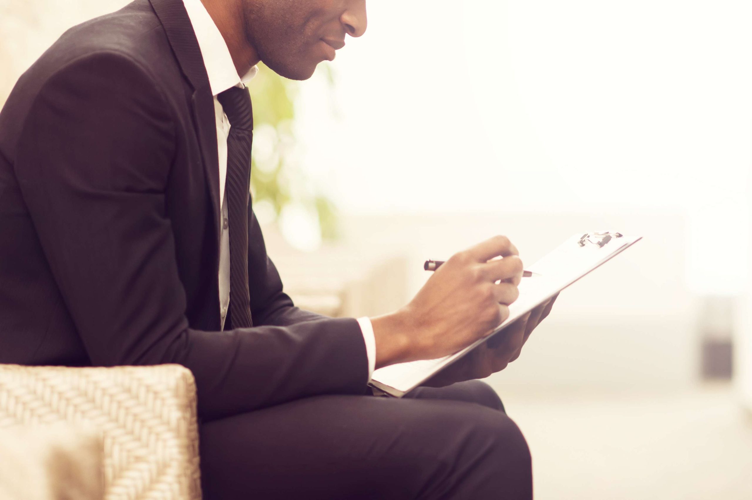 Making some business notes. Side view close-up image of cheerful young African businessman writing something in his note pad and smiling while sitting on the chair